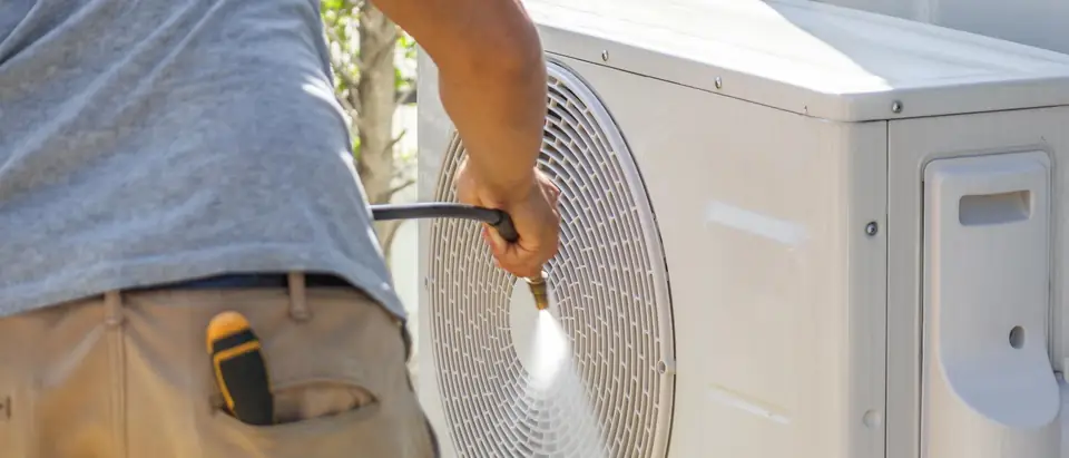A person seen from behind rinsing the fan grille of an outdoor air conditioner unit with a spray of water from a hose