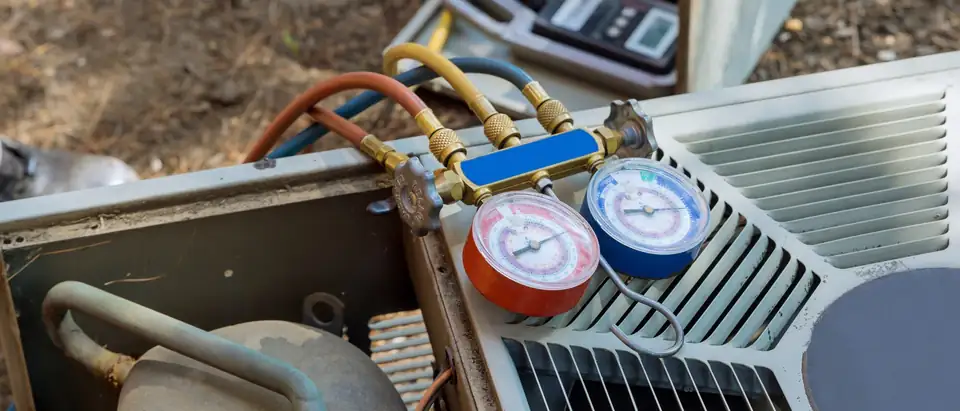 A set of red and blue refrigerant gauges with its hoses resting on top of an outdoor condenser unit whose side panel is off, showing the compressor