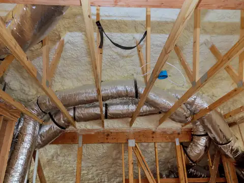 Silver insulated flexible ducts strapped along the roof trusses of an attic, with pale spray foam insulation behind them
