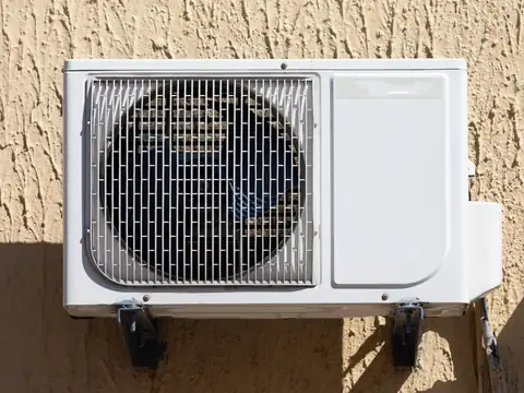 A white outdoor air conditioner unit on brackets against a rough tan stucco wall in bright sun, its fan visible behind the grille
