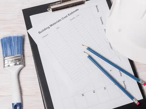 A blank cost estimate form on a black clipboard with two pencils lying on it, beside a white hard hat and a paintbrush