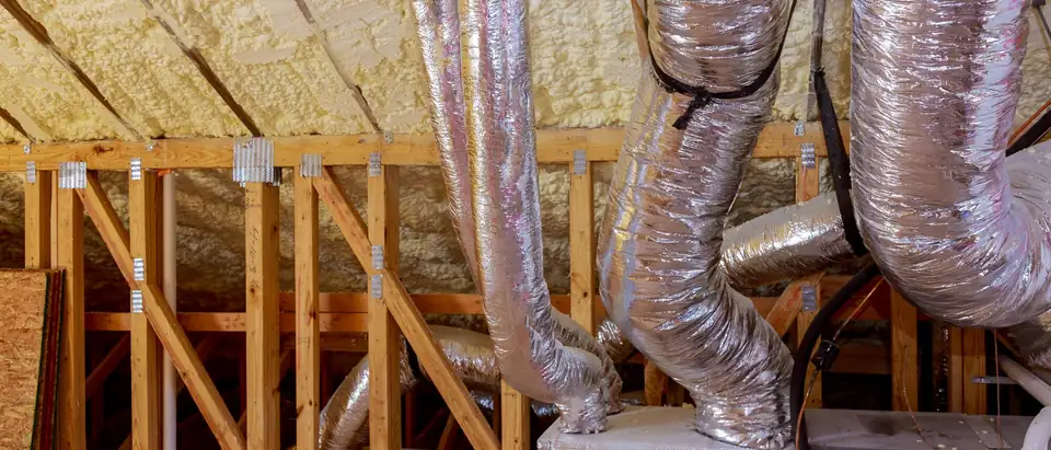 Silver insulated flexible ducts dropping from the rafters into a plenum box beside an air handler in the attic of a house under construction