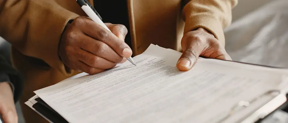 A hand holding a pen over the pages of a printed agreement on a clipboard, with the other hand steadying the paper