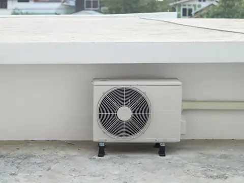 A clean cream-colored outdoor air conditioner unit standing on rubber feet on a concrete slab against a white wall